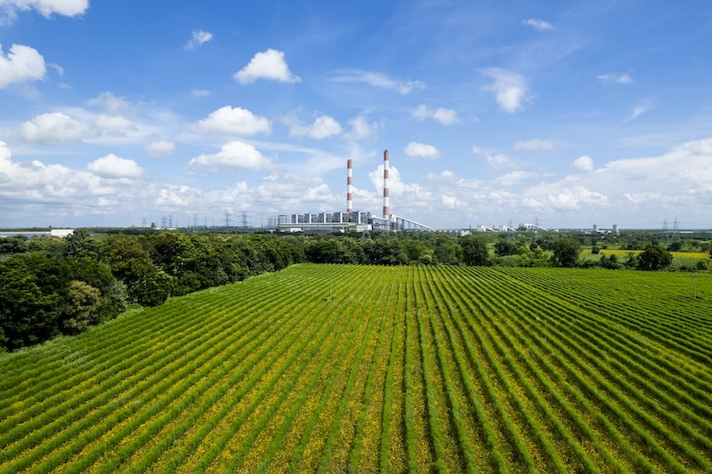 Blick auf Industrieanlage hinter grüner Feldlandschaft – Symbol für nachhaltiges Umweltmanagement der Analyze HSE GmbH